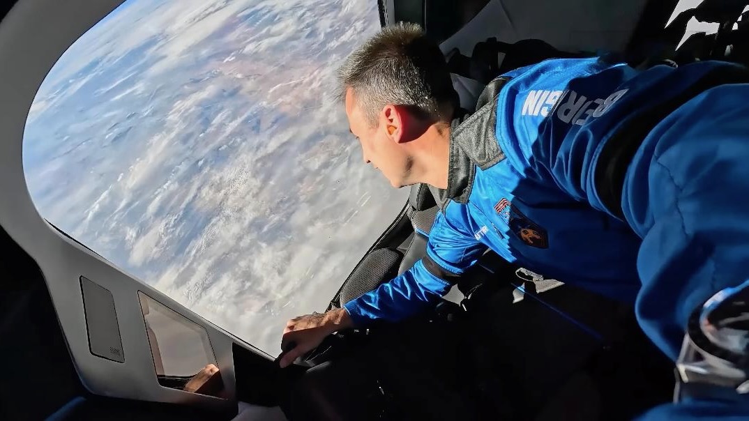 a male astronaut in a blue flight suit gazes out a large spacecraft window at earth below