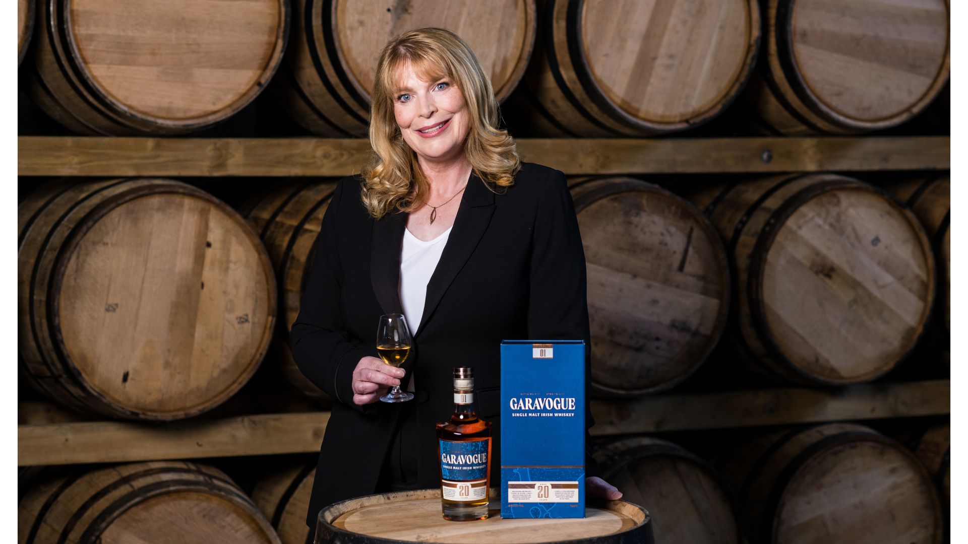 An image of Helen Mullholland, master blender at Hawk's Rock Distillery. She is standing in front of three rows of barrels. In front of her is the bottled Garavogue whiskey with the bottle box next to it on top of an upright whiskey barrel. She is holding a glass of Garavogue whiskey in a small fluted tasting glass