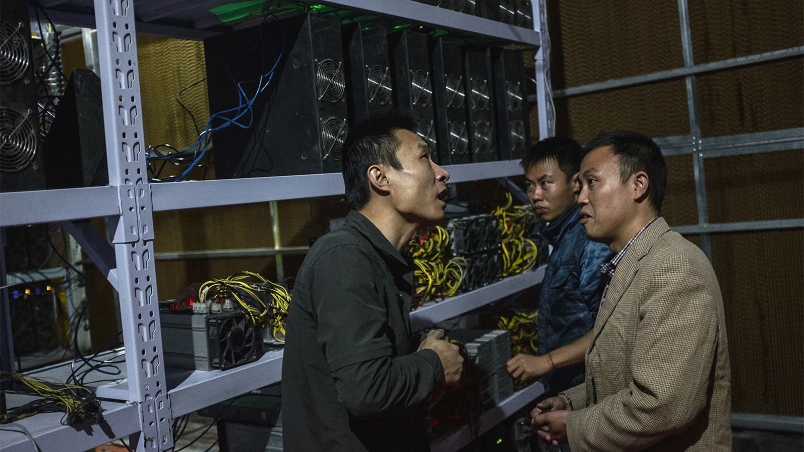 Three Men In China Speaking In Front Of A Rack Of Computers Mining Cryptocurrencies