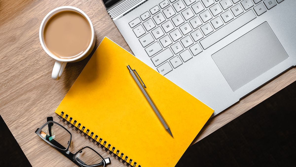 A laptop, yellow notebook, pen, cup of tea and pair of glasses laid out on a desk