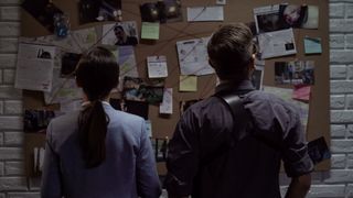 One female detective and a male detective looking at a bulletin board covered with images and red lines.