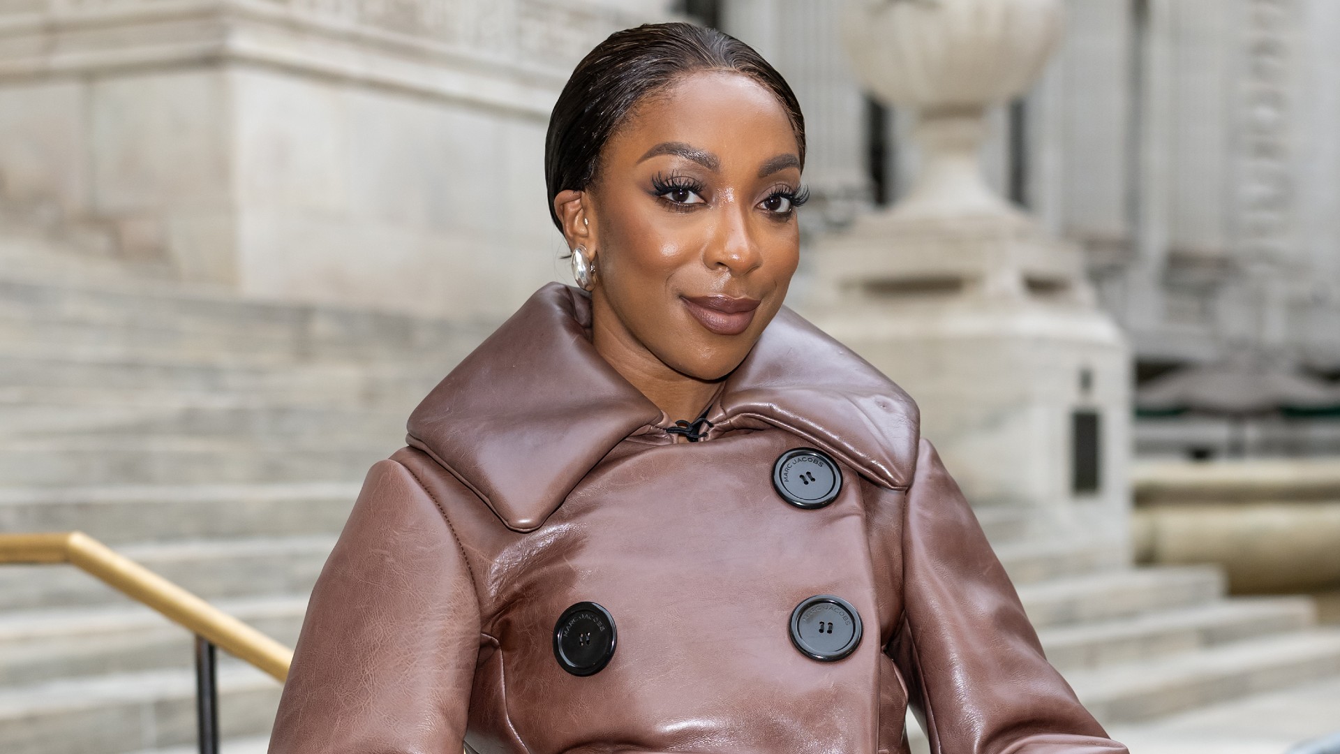 a black woman with dark hair pulled back into a bun posing in front of steps in a brown coat with black buttons