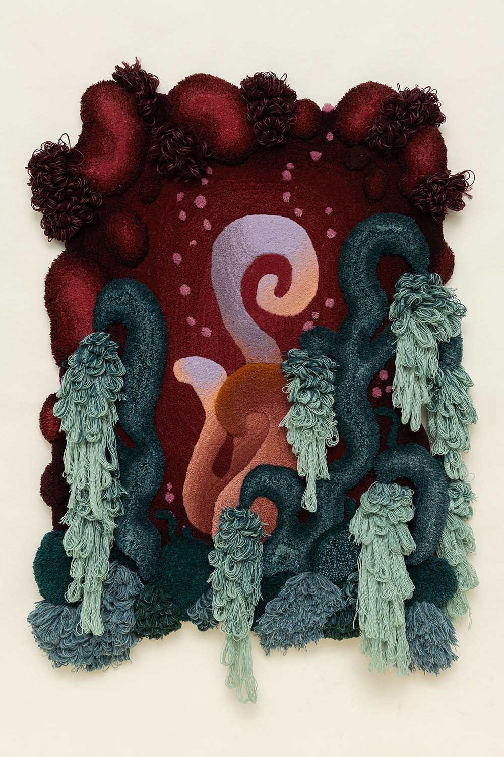 A tufted, woven textile creation hanging on a cream wall, with water-like segments mixed with dark red backgrounds and hems in an abstract composition.