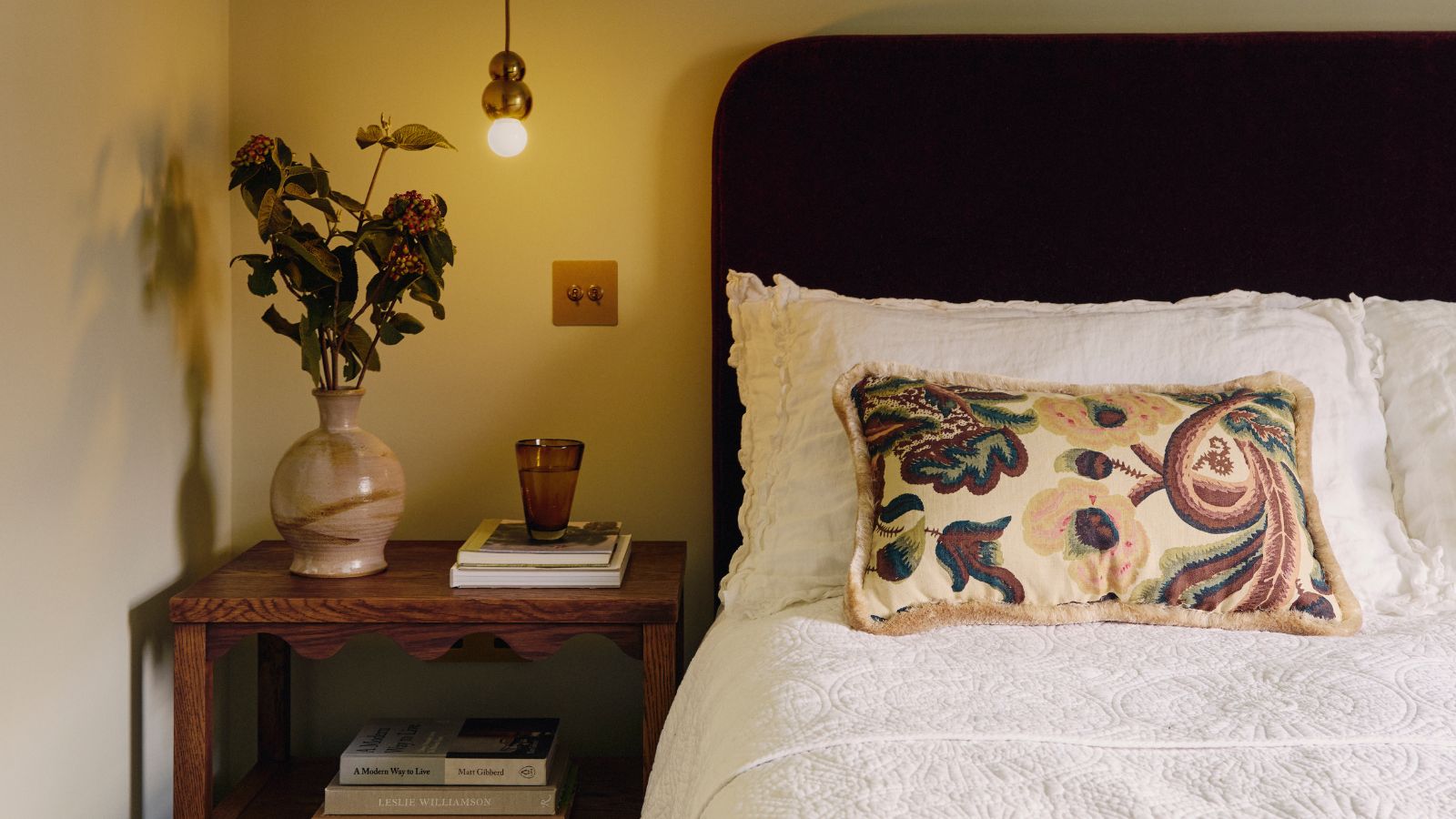 Bedroom with white bedding and dark headboard and a wooden vintage bedside table with a marble vase of fall foliage