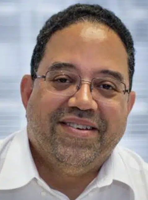 a black man with short dark hair wearing glasses and a white shirt smiling at the camera