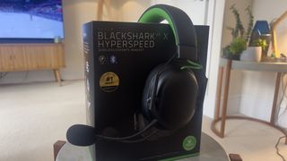 The Razer Blackshark V3 X gaming headset on a table. A living room is visible in the background.