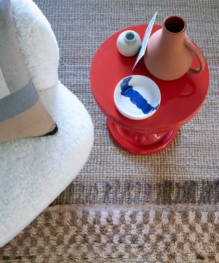 Close up of brown and beige checkered rug on beige textured carpet, right red round side table, ornaments on table, corner of cream lounge chair with cushion