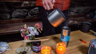 An MSR Titan Kettle is pouring water into a flask on a crowded bothy table.