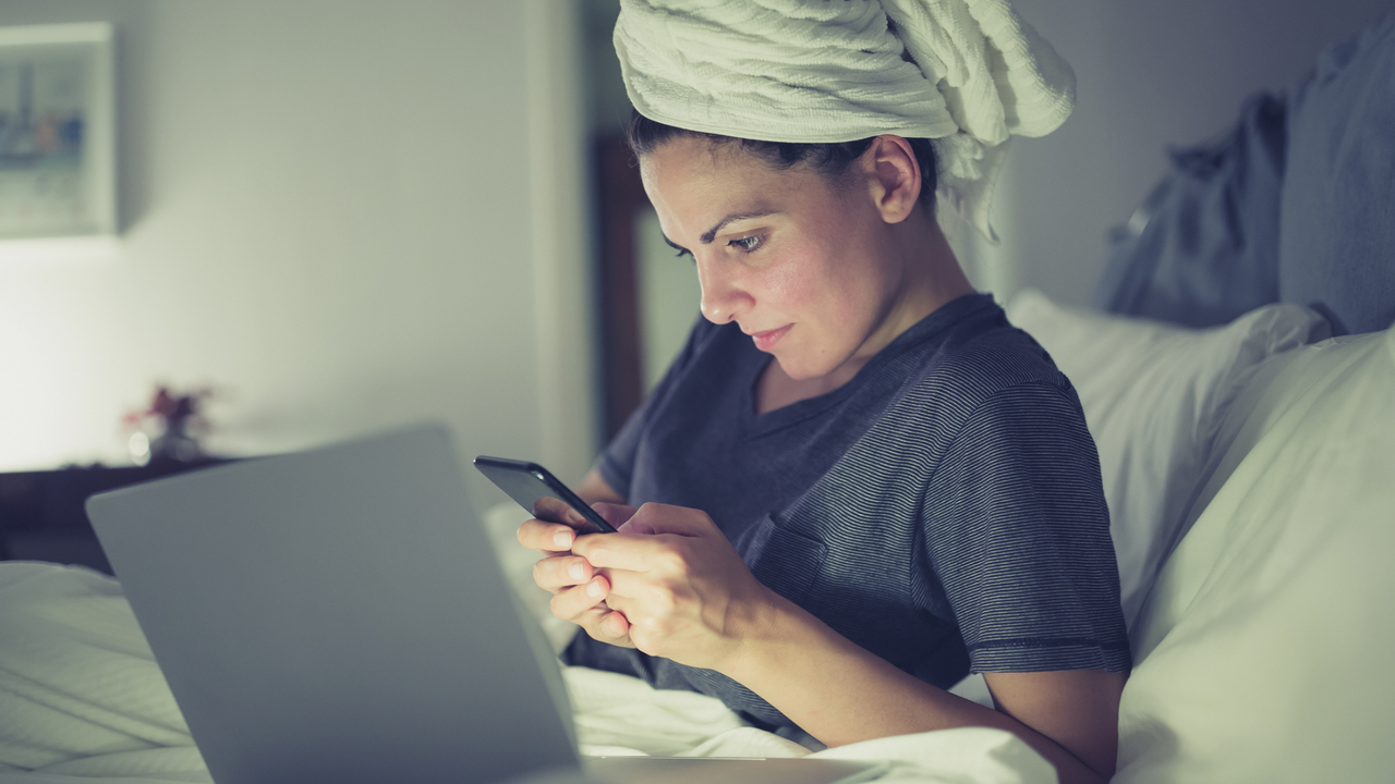 Woman working late at home using laptop and mobile phone. She is lying in bed with her hair wrapped in a towel.