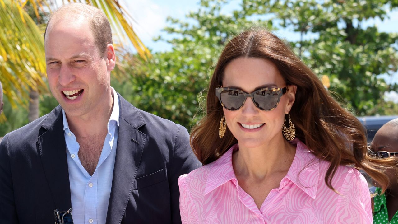 Prince William wearing a blue open necked shirt with a navy blazer standing outside next to Kate Middleton in a pink dress and gold hoop earrings with sunglasses