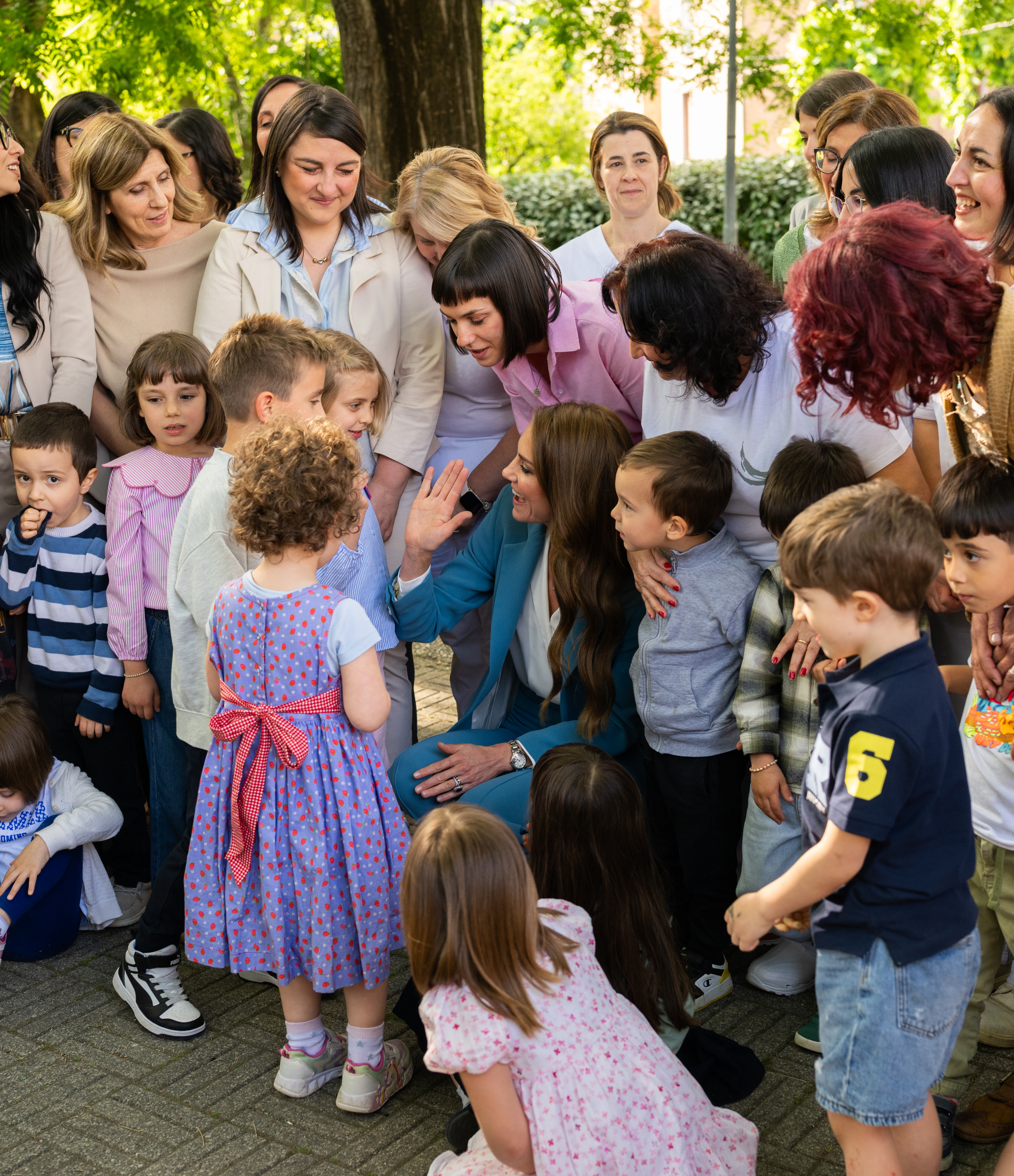 Princess Kate wearing a blue suit, kneeling down talking to kids and teachers in Italy