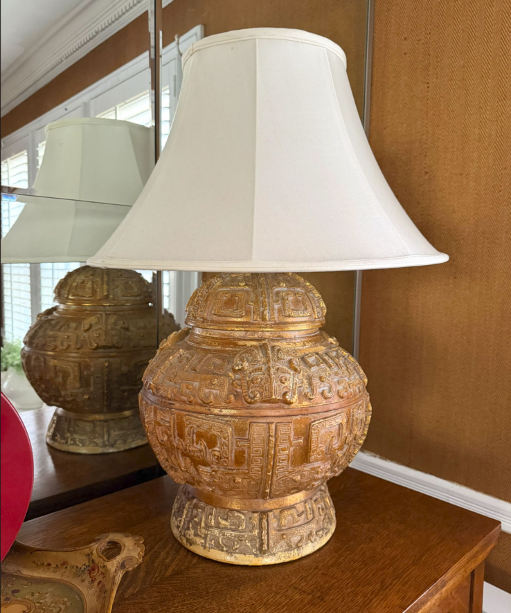Large lamp with white shade, antique golden base with intricate carving, placed on top of a wooden side table, mirorr in background