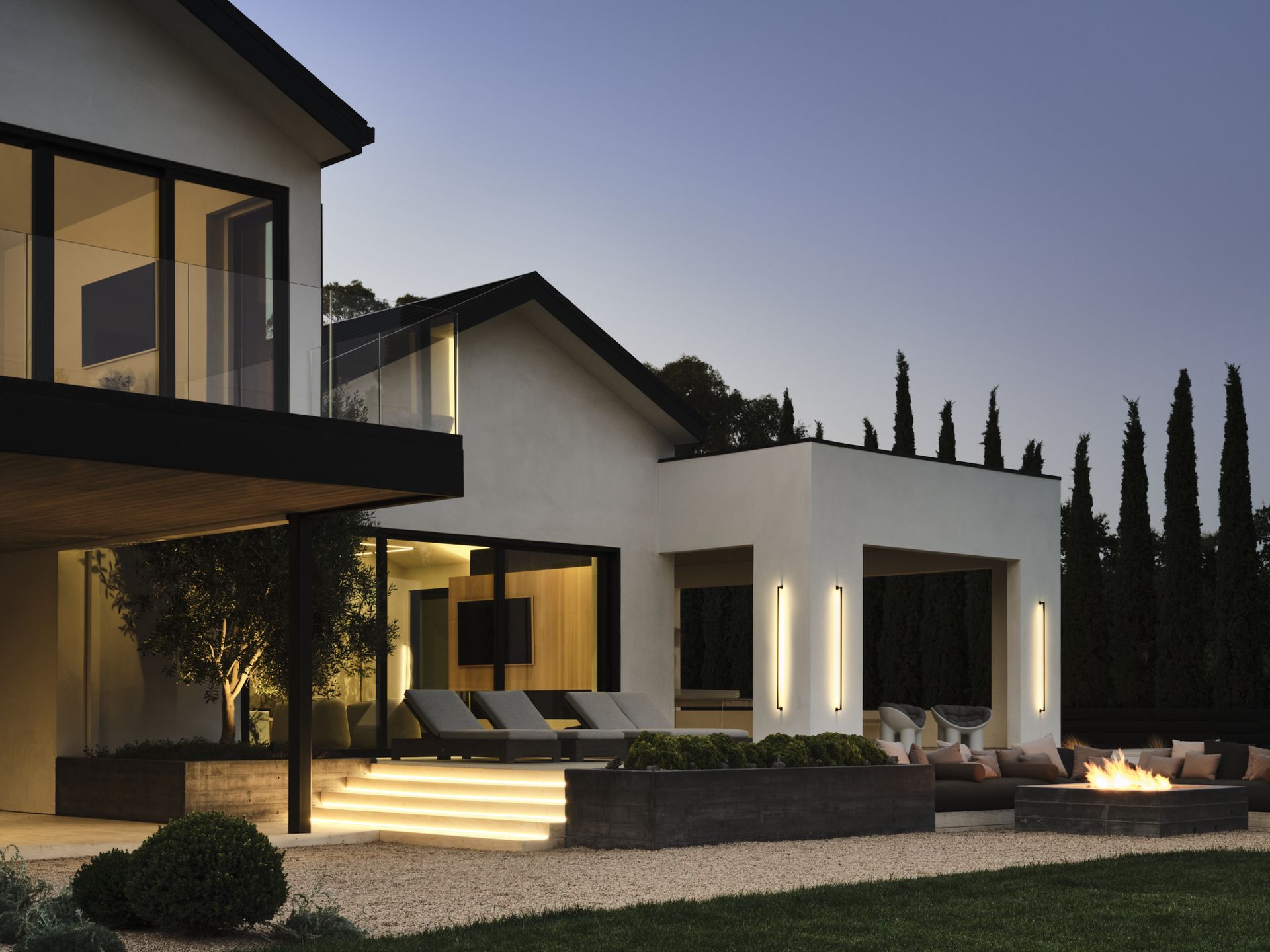 An outdoor area of a modern home with large black framed windows complete with a lush garden and steps illuminated with recessed lights and a lit fire pit among outdoor seating at dusk