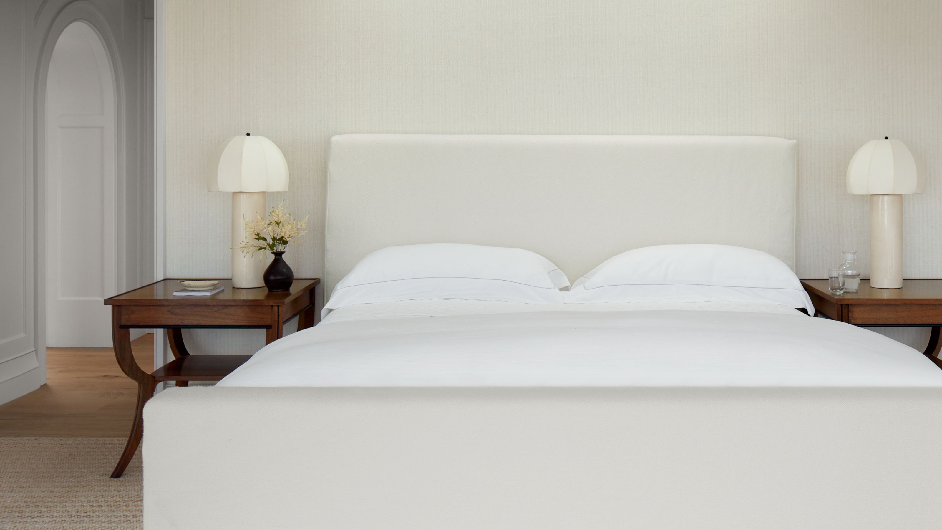A serene, minimalist bedroom with an upholstered cream headboard and crisp white bedding. Two dark wood nightstands hold large sculptural lamps with pleated shades, set against a subtle textured wallpaper.