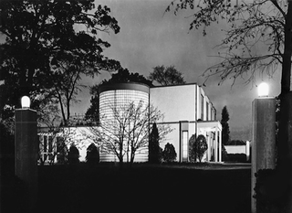 Brunning House (1936), black and white night image, driveway entrance stone pillars lit up, surrounding trees and landscape, building glows white against the night sky