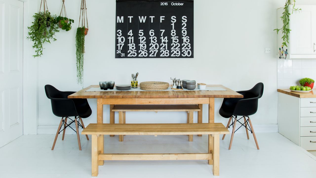 white dining room with hanging plants, big wall hanging, blond wood table with matching benches, black Eames style chairs