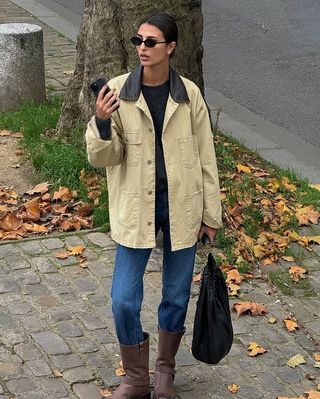 French Fashion Trends 2025: @salome.mory wears a barn jacket with blue jeans and brown boots whilst standing on a leafy street