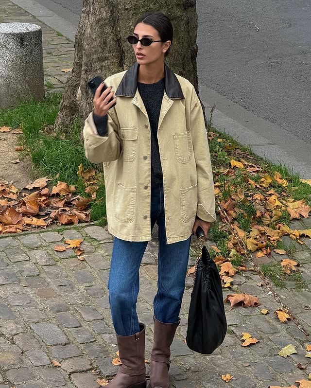 French Fashion Trends 2025: @salome.mory wears a barn jacket with blue jeans and brown boots whilst standing on a leafy street