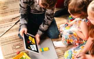 Young boy with vision impairment, has a pale complexion and short brown hair, and wears black framed glasses, shows a tablet displaying an online LEGO Braille Bricks PlayStarter activity to his two sisters. The youngest wears pink framed glasses. Both have pale complexions and light brown hair.