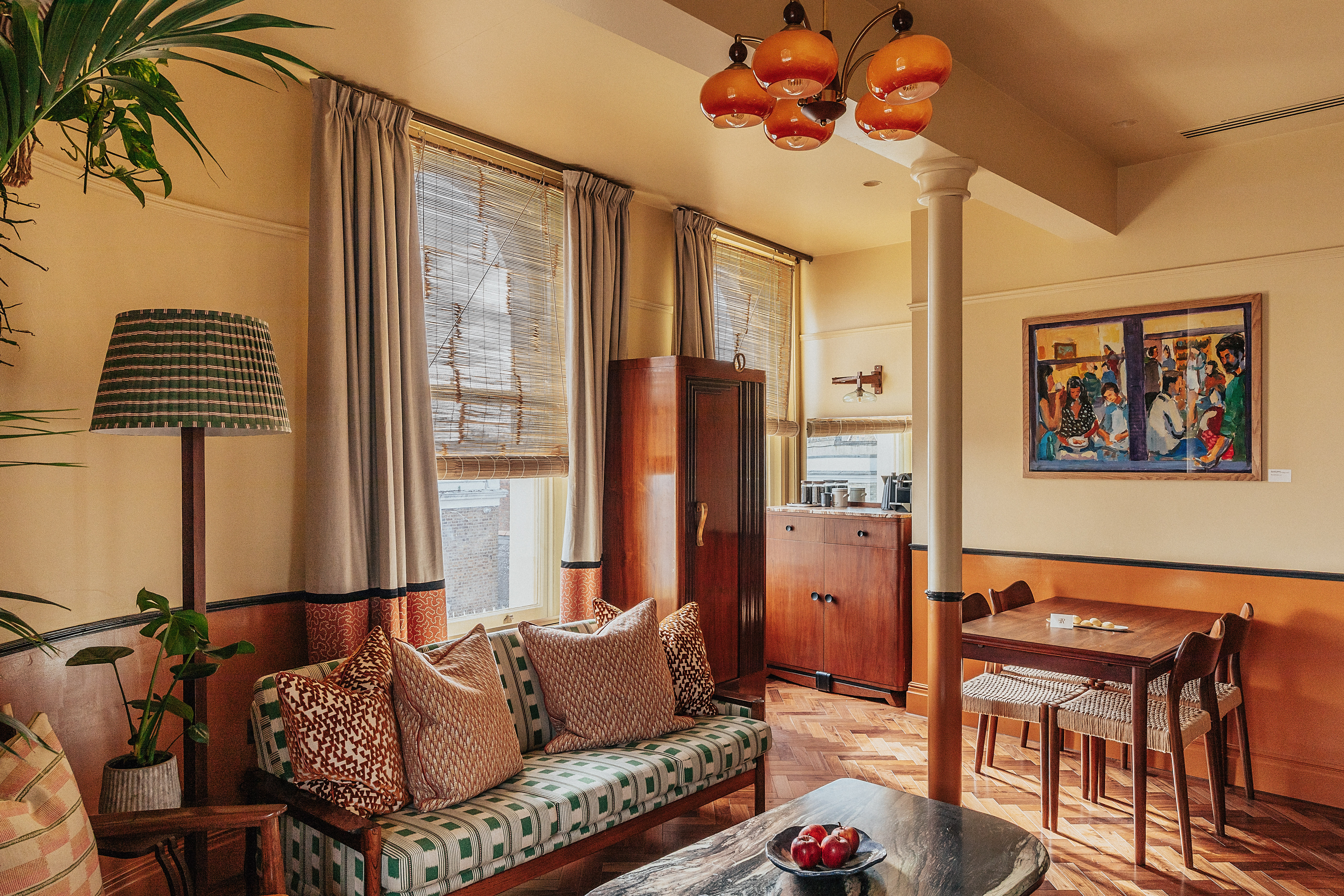A vibrantly decorated hotel suite inspired by Bombay residences features buttery or tile blue walls adorned with original portraiture artworks, bespoke retro-fueled furnishings in wood, leather, and patterned fabric, and thriving plants.