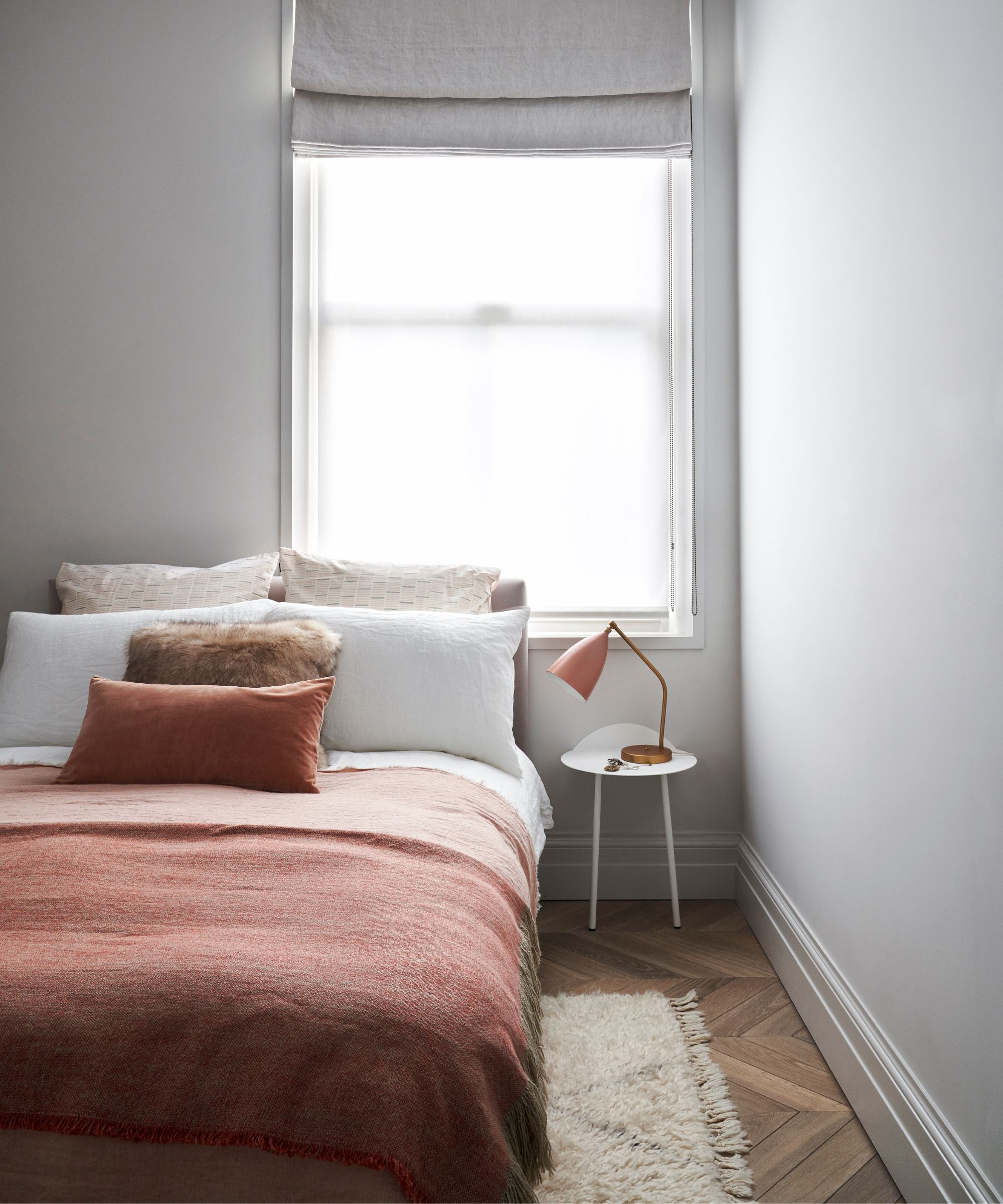 A bright white bedroom with brown wooden flooring, and a bed with burnt orange colored bedding. Underneath the bed is a white fluffy rug, and on the white bedside table is an orange table lamp.