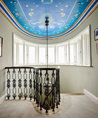 Large landing space with stand out painted ceiling, blue patterned design, large staircase with black railings, beige carpet, gray painted walls, looking out to curved windows