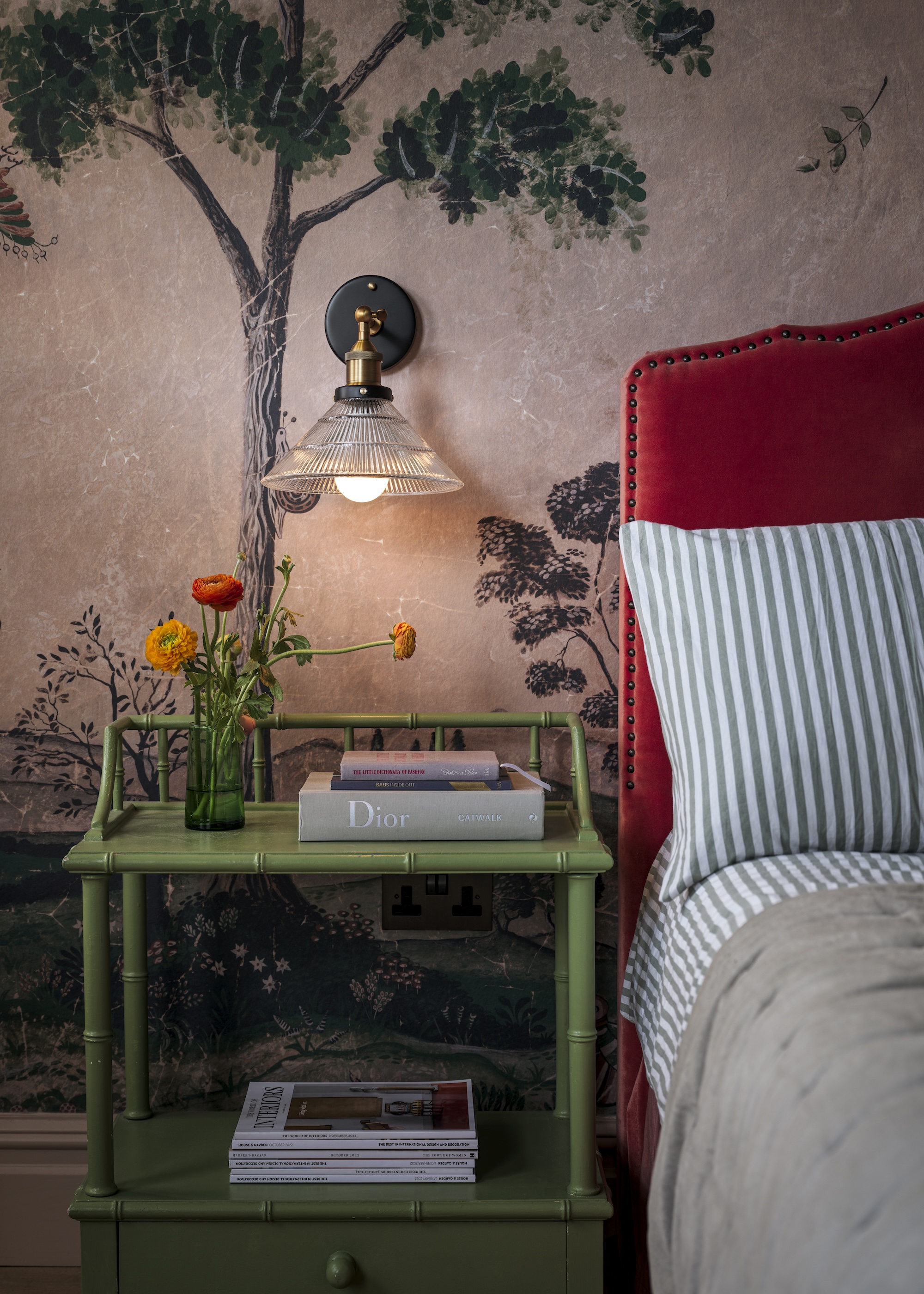 A bedroom with a green bedside table and a wall lighting hanging above the table. On the bedside table are some books and magazines as well as a glass with flowers inside of it.