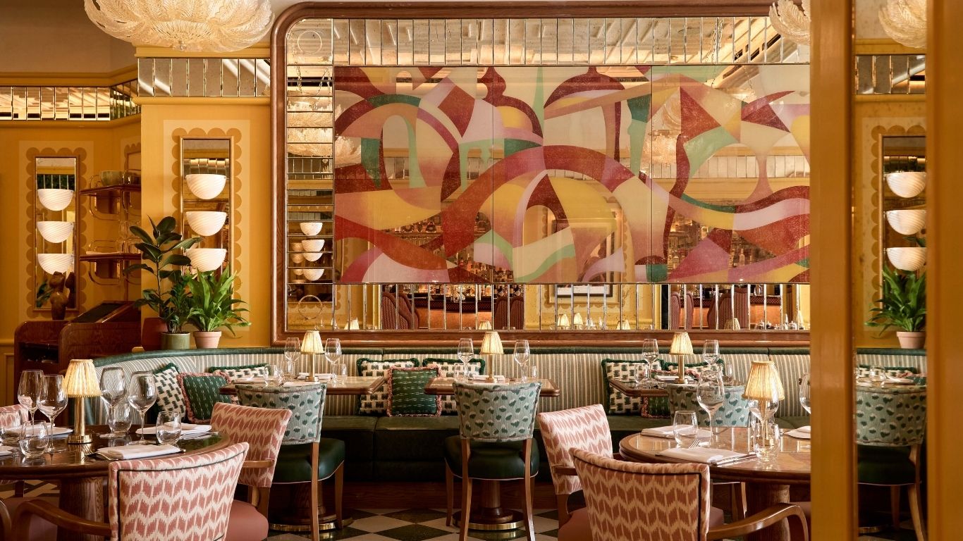 A huge mirror painted in abstract pastel shapes overlooks the green and pink upholstered seating area of the Cicchetti Piccadilly restaurant