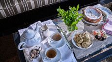 Teapot and cups and saucers laid on a silver tray with a plate of biscuits, a cake, some mint leaves on a rustic chest