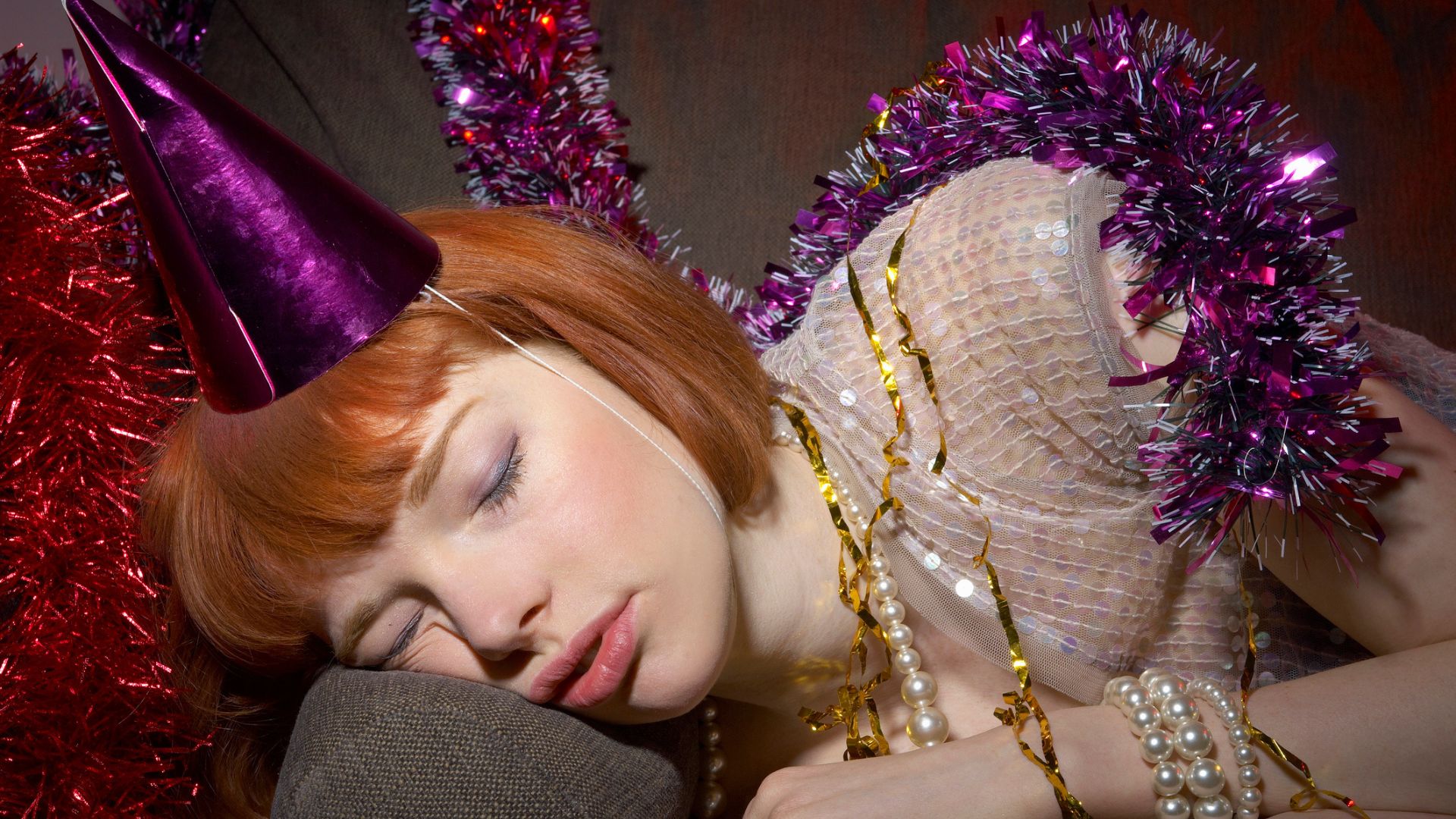 A woman with red hair wearing a glittery dress and a party hat asleep on a sofa