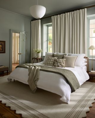 The scalloped beige rug in a cozy neutral bedroom. There is a bed with white and gray-green bedding