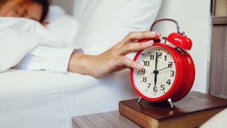 A woman lifts her arm out from under her white duvet in order to turn off her alarm clock