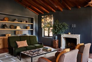 A dark living room with wooden beams and built-in cabinetry with a green sofa facing two light brown armchairs with a rectangular coffee table in the middle in front of the white fireplace.