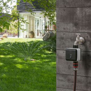 smart watering system attached to garden tap which is attached to the exterior wall of a house with the garden lawn and house in the background