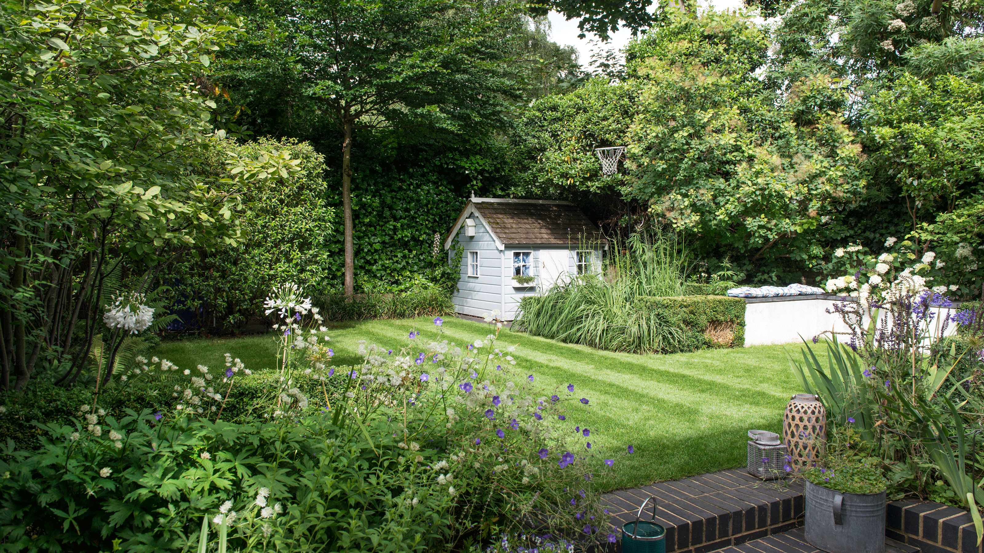 A garden with a tidy lawn and flowerbeds, wooden deck, raised bed edging, and plank path, and grey fence and sun loungers