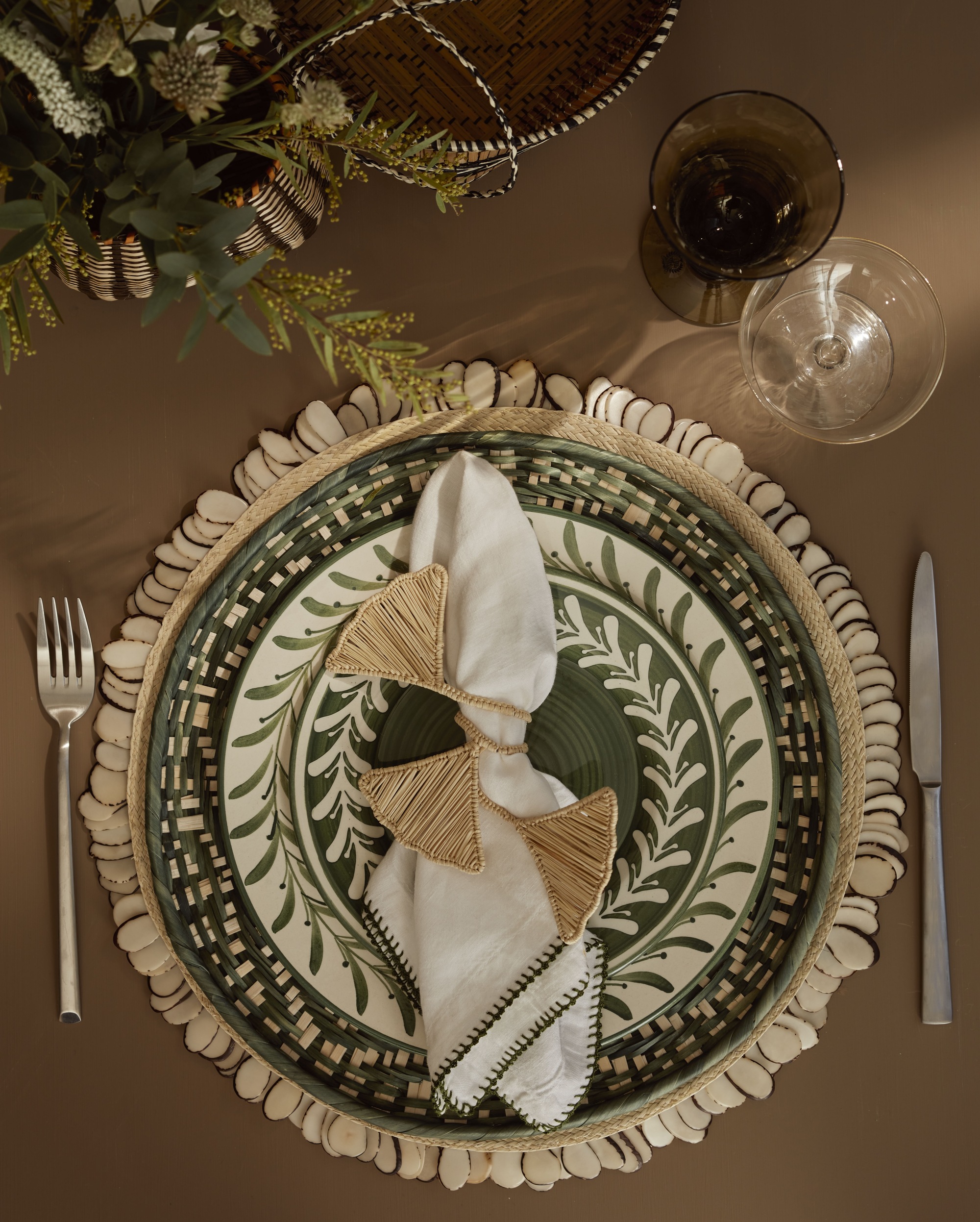 table setting with brown tablecloth, with raffia placemat, green patterned plates, napkins and napkin ring, and silver cutlery, and wine glasses, with floral centerpiece