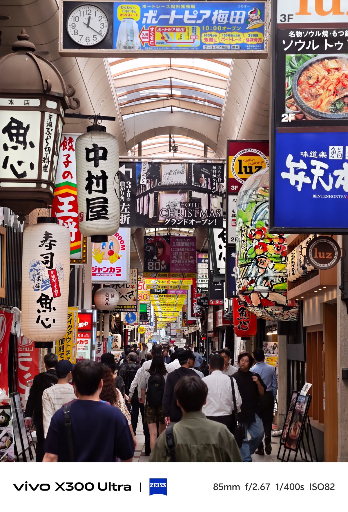 A rear view of pedestrians walking down a covered market alley packed with large hanging lanterns and signs.