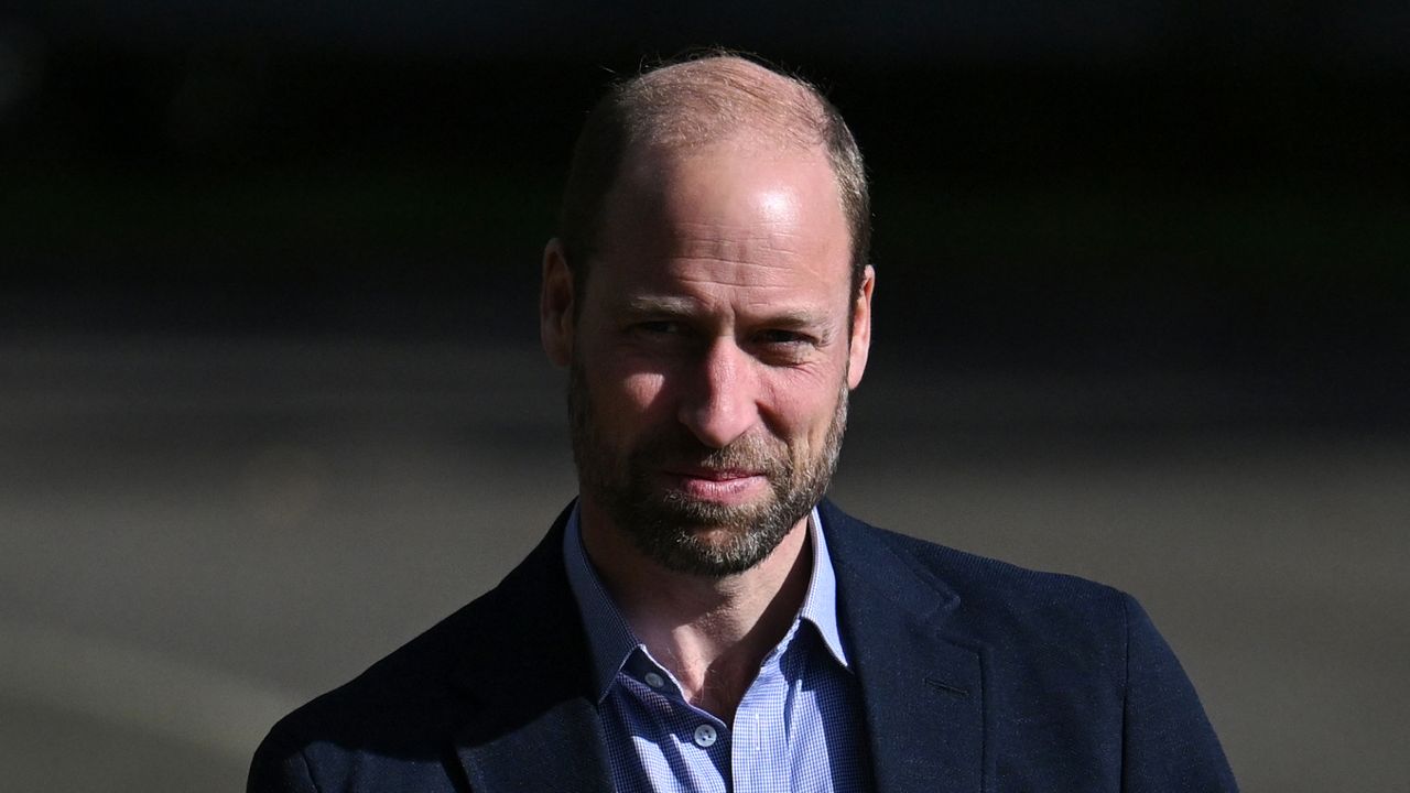 Prince William has a beard and smiles while wearing a casual suit jacket and unbuttoned blue shirt