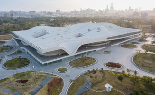 National Kaohsiung Center for the Arts, Taiwan, by Mecanoo. A large structure with a wavy roof which is surrounded by walkways and plant areas.