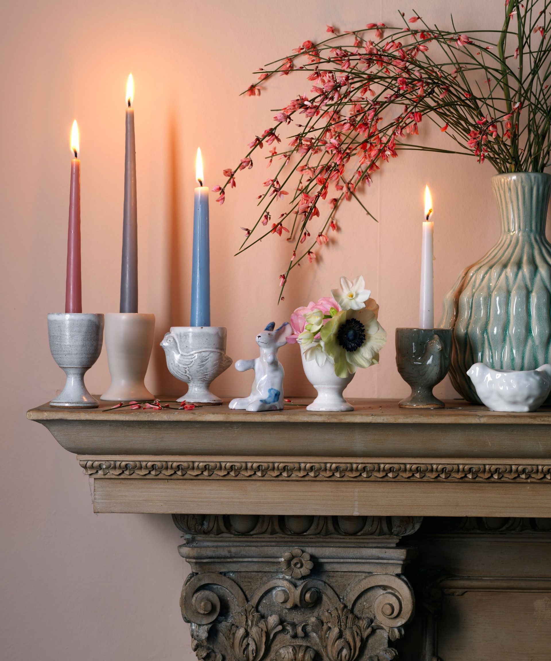 A pink wall with a brown wooden mantelpiece, with tall, thin pillar candles in various colors in eclectic holders. There is also a large green vase with red leafy stems.