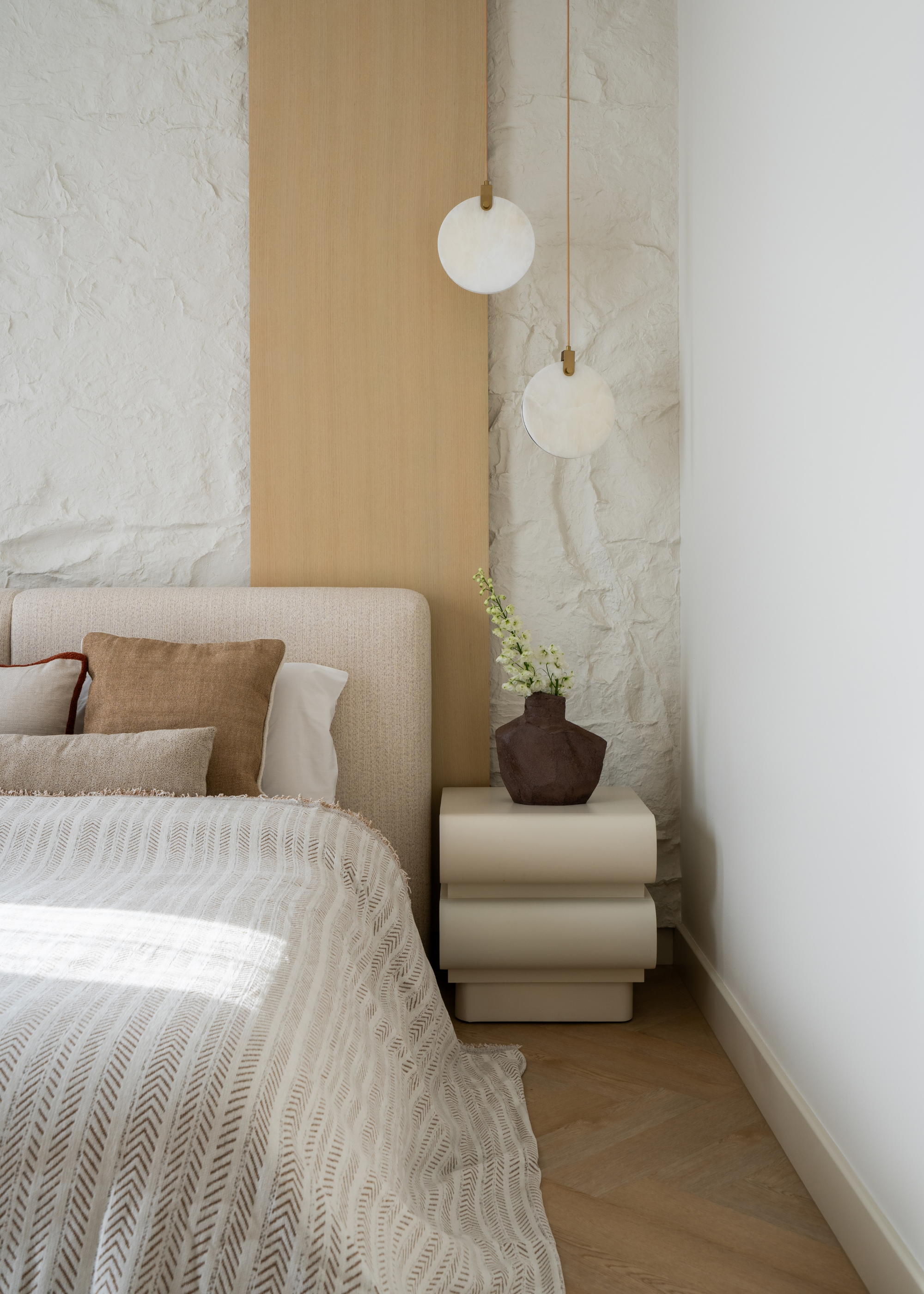 A minimalist bedroom with textured walls, a cream side table, a vase of flowers, a pendant light, and a bed with cushions