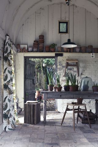A rustic potting shed with a wooden potting table, fern print drapes and stone flooring illustrating she shed ideas inside.