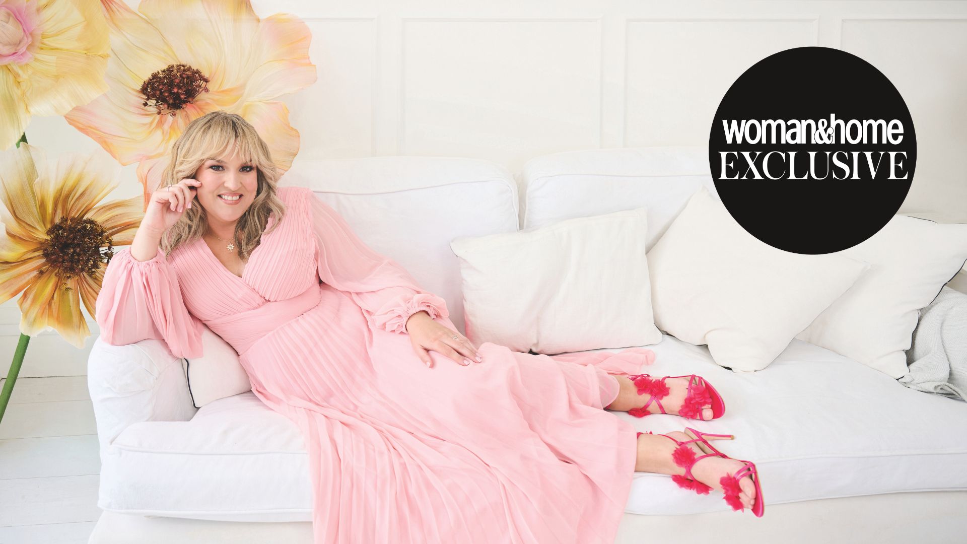 Sara Davies in a pink dress and red shoes on a sofa 