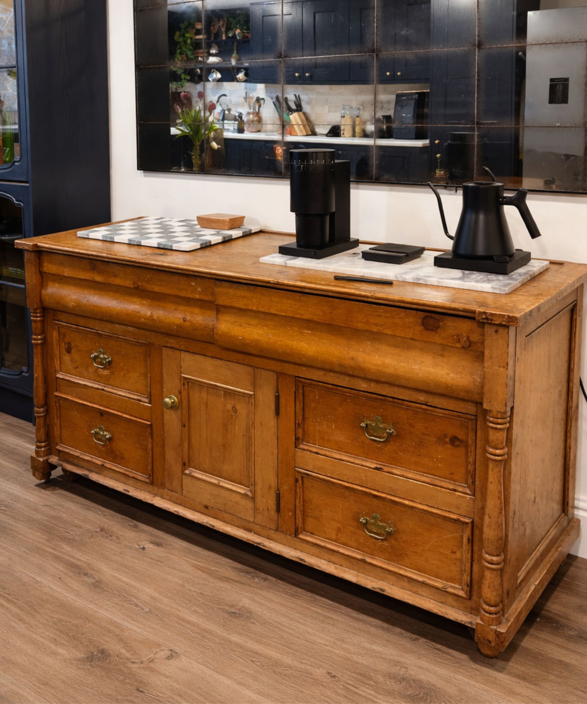 Vintage wooden sideboard with brass handles beneath a large grid mirror, styled with a black coffee grinder and kettle on marble trays in warm light.