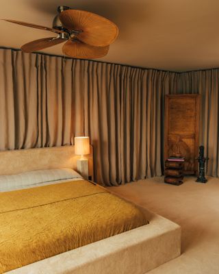 A beige, brown, and yellow bedroom. The bed is a textured velvet bed with mustard yellow bedding. Along the back wall is a thick line of taupe curtains.