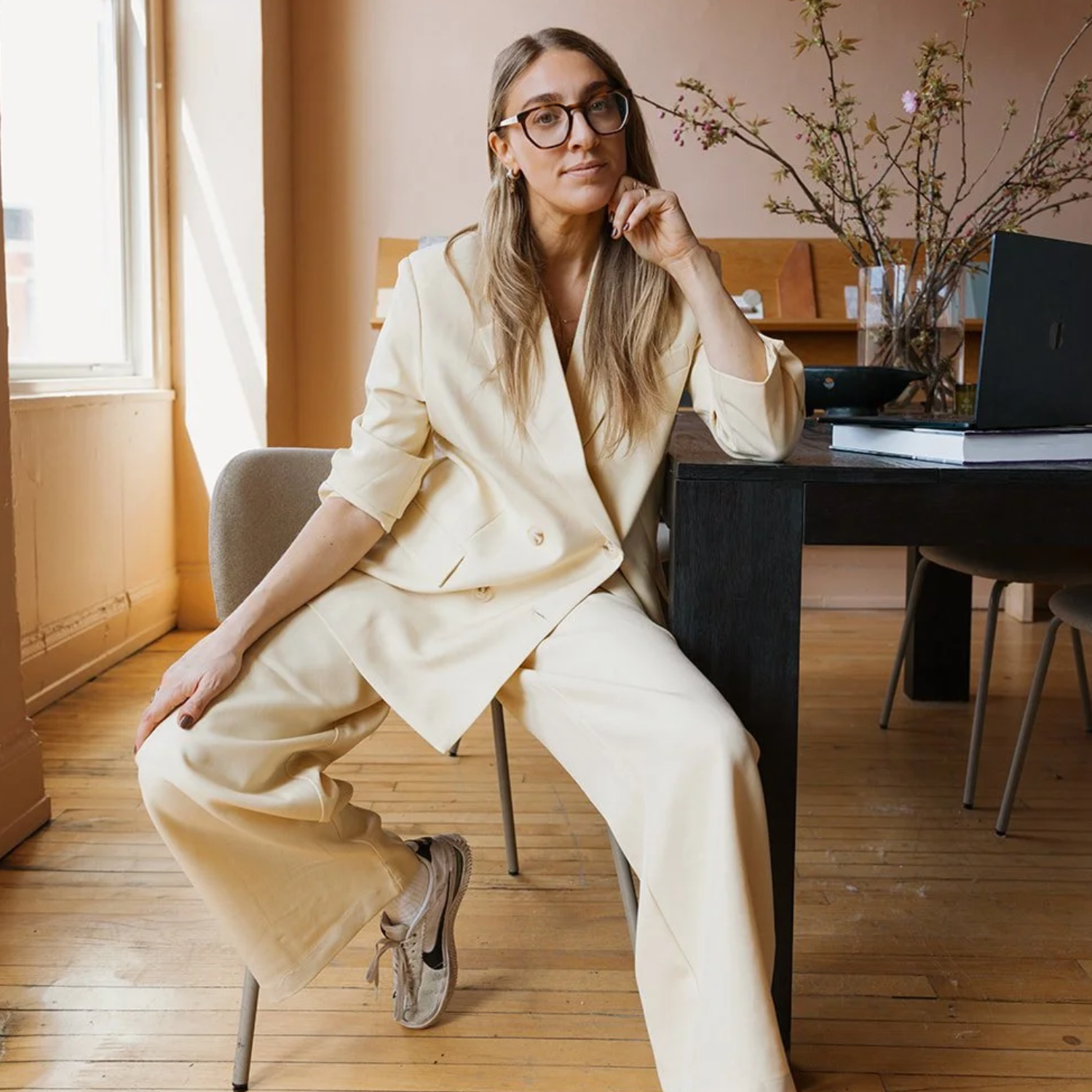 A woman in an ivory suit is posing on a chair by a black table