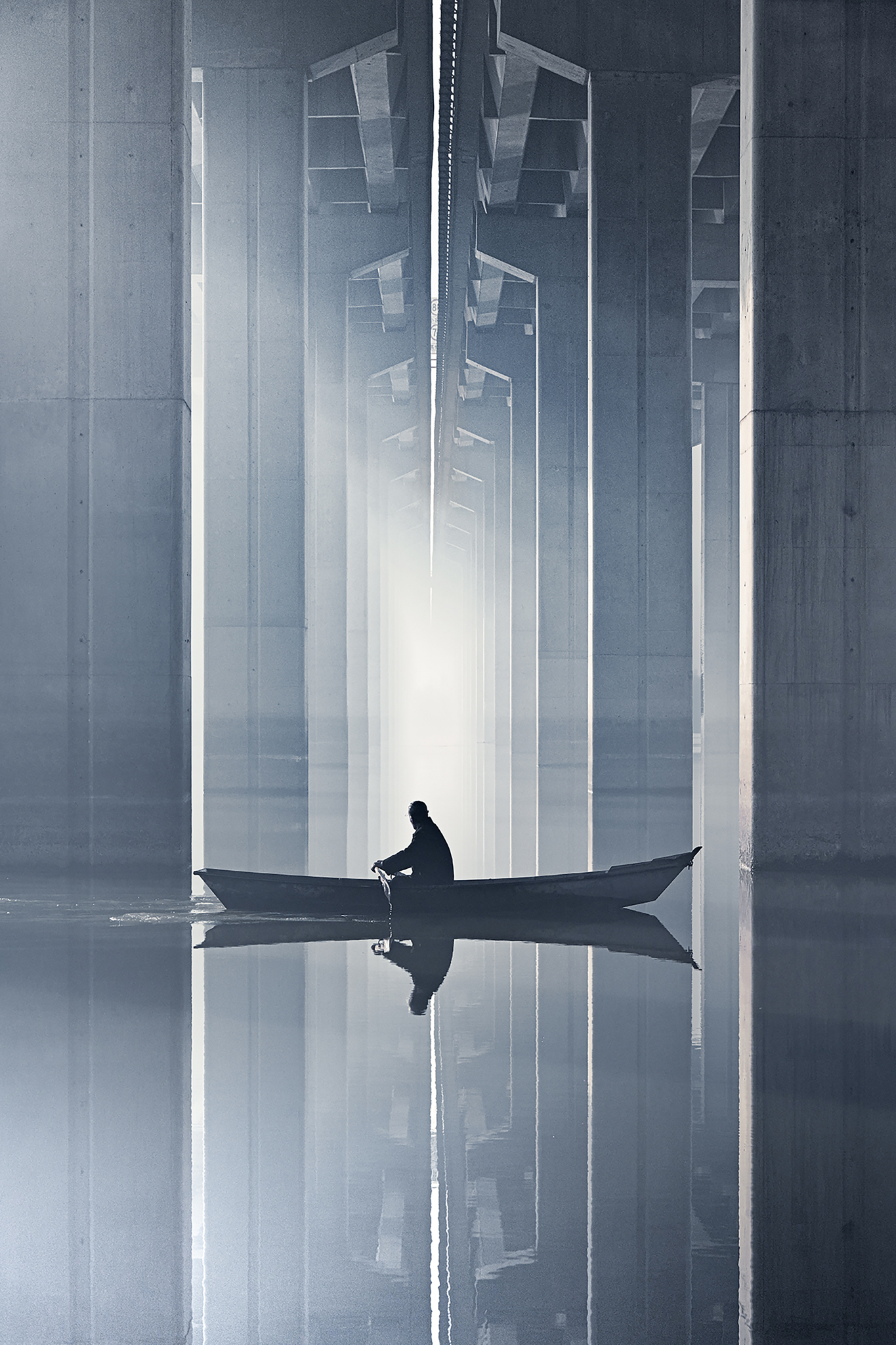 A person rows a small boat through calm waters under a modern bridge, with towering concrete beams