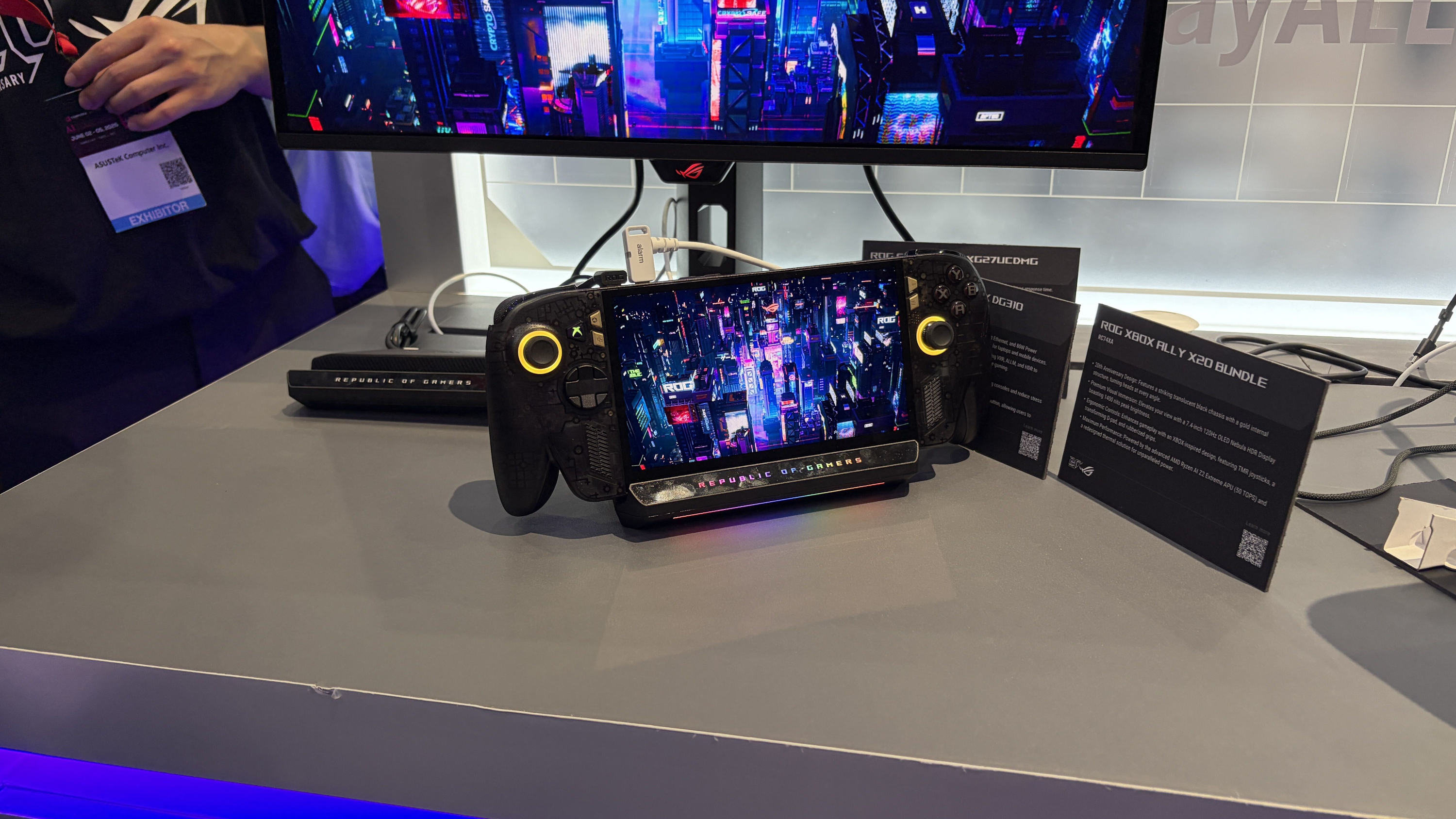An XBOX Ally X20 handheld gaming device with a vibrant cityscape on the screen, mounted on a display booth with "Republic of Gamers" illuminated below it.