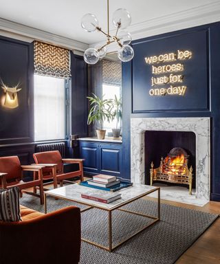 Living room with dark blue panelled walls with a lit fire and neon sign, wooden flooring with a grey rug and square marble topped coffee table and marble framed fireplace.