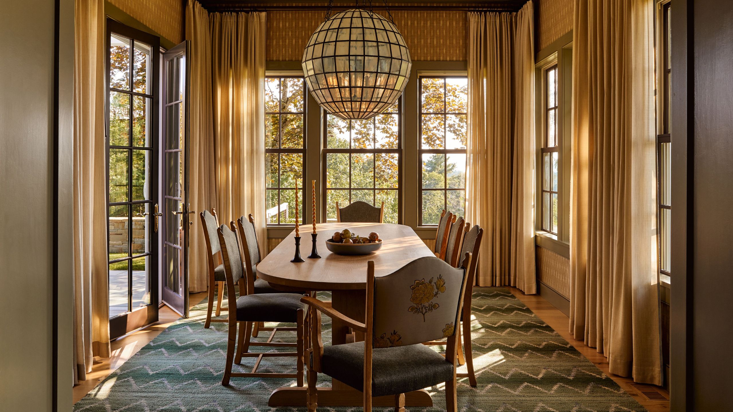 a dining room surrounded by windows and doors with a large curved wooden table, ornate dining chairs with floral fabric, large glass globe pendant and blue zigzag rug